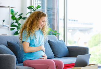 Obraz premium Caucasian happy pretty long curly hair healthy pregnancy mother in casual outfit sitting smiling on cozy sofa touching rubbing tummy belly watching online streaming via laptop computer in living room