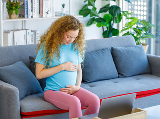 Obraz premium Caucasian happy pretty long curly hair healthy pregnancy mother in casual outfit sitting smiling on cozy sofa touching rubbing tummy belly watching online streaming via laptop computer in living room