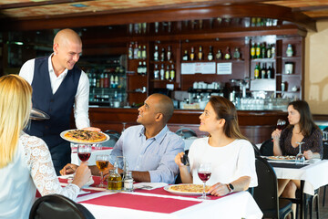 Friendly smiling waiter bringing ordered pizzas to guests of cozy italian restaurant