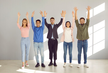 Portrait of cheerful young people standing in row with arms raised and smiling in front of camera. Happy different men and women in casual clothes standing in empty room with sunlight.