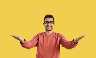 You did it. Congratulations. Happy cheerful positive young ethnic man in eyeglasses standing against yellow color background looking at camera, smiling and spreading his arms wide open for a warm hug - obrazy, fototapety, plakaty