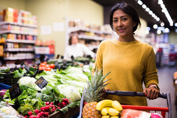 Portrait of smiling latin american woman with cart of fresh grocery, enjoying shopping in store