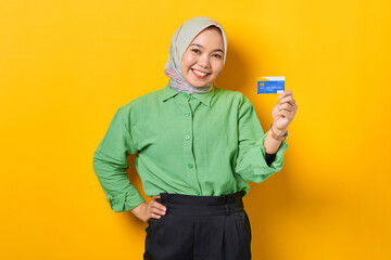 Smiling young Asian woman in a green shirt showing credit card on yellow background