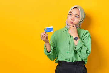 Pensive young Asian woman in a green shirt holding credit card and looking away on yellow background