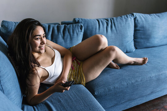 young latin woman lying relaxing on sofa in living room and watching TV at home in Mexico Latin America	