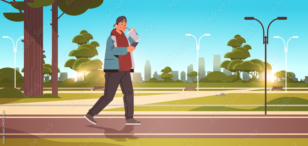 Poster man drinking coffee with straw and using smartphone while walking in urban park social media communi