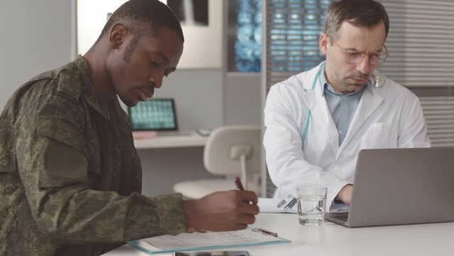Waist up slowmo of African American soldier in military uniform filling out medical form during his appointment at modern doctors office, Caucasian male radiologist sitting nearby