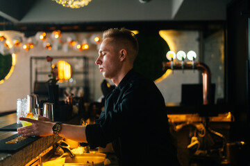 Side view of professional young barman giving ready cocktail to customer standing behind bar counter on blurred background of modern nightclub with dark interior, selective focus.