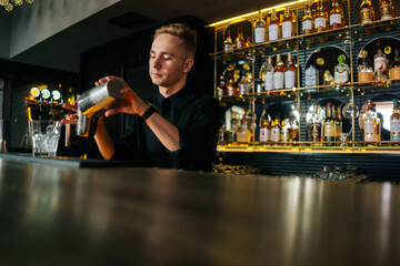 Low-angle view of handsome bartender preparing refreshing alcoholic cocktail standing behind bar counter in modern dark nightclub, on blurred background of shelves with different alcoholic drinks.