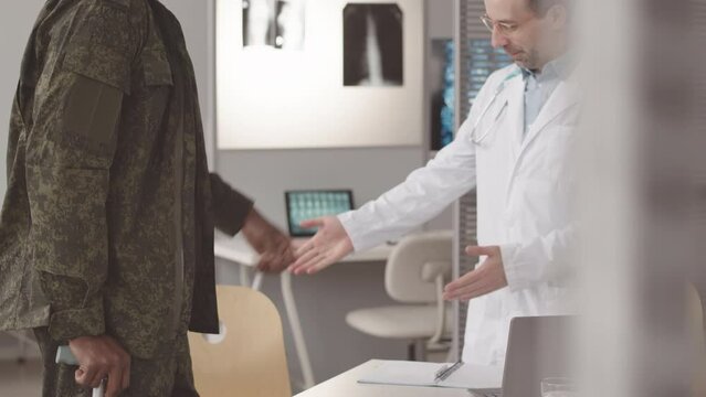 Medium slowmo of young African American army soldier in military uniform limping and leaning on stick and Caucasian male doctor having medical appointment in clinic, greeting each other with fist bump