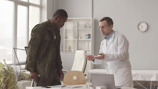 Medium slowmo of Caucasian male doctor giving prescription to African American soldier leaning on walking stick, having medical appointment at modern doctors office