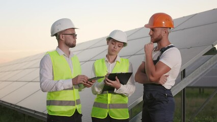 Two inspectors in white helmets using tablet and clipboard during conversation with competent technician at solar station. Maintenance and green energy production.