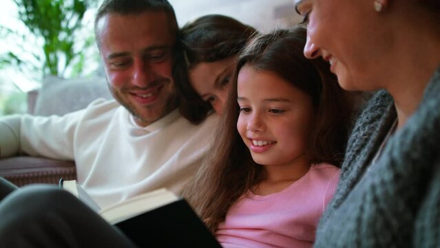 Two happy sisters with mother and father sitting on floor hugging and reading book at home