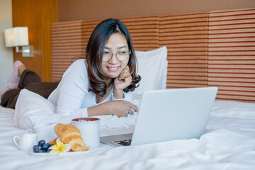 Young Asian women smiling happily at freelance work, working on a notebook while relaxing in bed with snacks and fruit in a hotel room. Vacation and relaxation