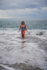 A plump woman in a bathing suit enters the water during the surf. Alone on the beach, Gray sky in the clouds, swimming in winter.