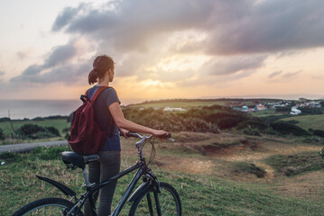 Fototapeta premium Young woman in the evening and watching the sunset while standing with her bicycle on a hill