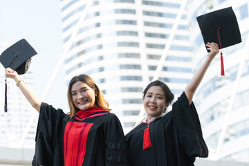 Two Asian young beautiful graduate female students with master and bachelor degree holding black board cap with red tassels up to the sky is smiling. Blur background of University building - obrazy, fototapety, plakaty