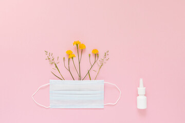 A set of wild flowers and herbs under a disposable protective mask and a mock-up of nasal spray on a light pink background. The concept of means of protection and treatment of seasonal allergies.