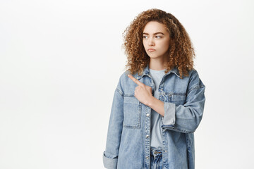 Portrait of angry, disappointed moody girl sulking, frowning upset, pointing finger left, standing over white background