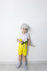A little boy in retro style with a yellow rose in his hand stands near a white wall.