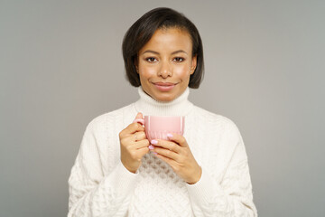 Happy carefree young african american woman smiling broadly, enjoying good coffee from mug. Attractive black female dressed in cozy warm knitted sweater drinking tea isolated on grey studio wall. 