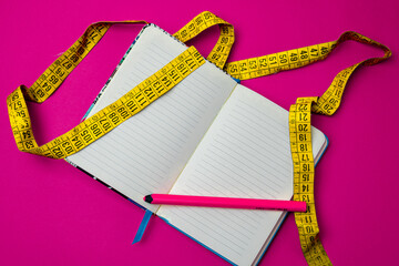 Close-up of a yellow tape measure rolled up on a diary with a pink pencil on a pink background. Concept of food care, weight problems and sports fitness.