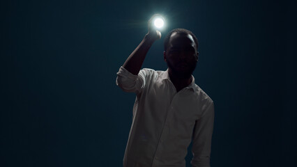 Young person sitting in the dark and using flashlight to search around, looking for evidence. Male detective taking risk and holding lantern to investigate and examine creepy sound. - obrazy, fototapety, plakaty