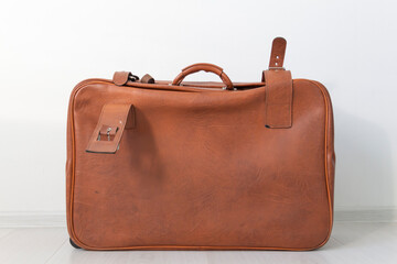 A brown, old, leathery, closed suitcase in retro style is located near the white wall.