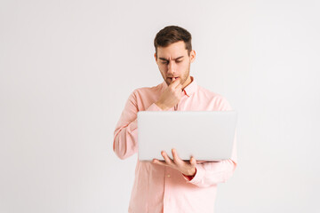 Portrait of serious pondering young man standing with laptop looking on screen with confused puzzled face on white isolated background. Studio shot of pensive male posing with pc looking at screen