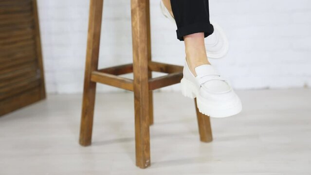 Presentation of white shoes with plain massive soles. Lady in modern footwear sits on the tabouret waving her feet. Close up.