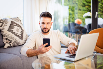 Fototapeta premium Serious faced man text messaging and using laptop while working from home