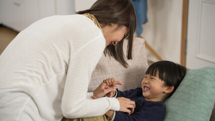 naughty Asian little boy making funny face expression to his mom as she is tickling his belly on the sofa in the living room at home