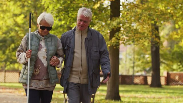 Elderly Caucasian man supports his blind wife while walking in the park disabled blind people support concept . High quality 4k footage