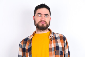 Fototapeta premium young caucasian man wearing plaid shirt over white background crosses eyes, puts lips, makes grimace with awkward expression has fun alone, plays fool.