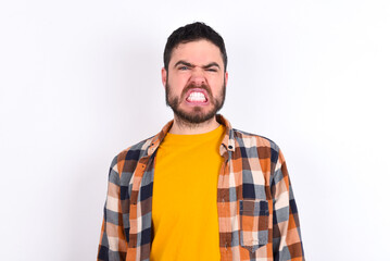 Fototapeta premium Mad crazy young caucasian man wearing plaid shirt over white background clenches teeth angrily, being annoyed with coming noise. Negative feeling concept.