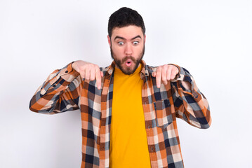 Amazed young caucasian man wearing plaid shirt over white background points down with fore fingers,...