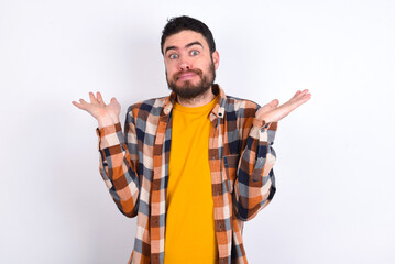 Puzzled and clueless young caucasian man wearing plaid shirt over white background with arms out, shrugging shoulders, saying: who cares, so what, I don't know. Negative human emotions.