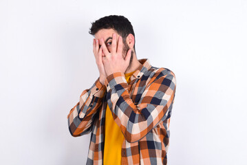 young caucasian man wearing plaid shirt over white background covering face with hands and peering out with one eye between fingers. Scared from something or someone.