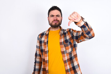 Discontent young caucasian man wearing plaid shirt over white background shows disapproval sign, keeps thumb down, expresses dislike, frowns face in discontent. Negative feelings.