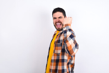 Fototapeta premium Overjoyed young caucasian man wearing plaid shirt over white background glad to receive good news, clenching fist and making winning gesture.