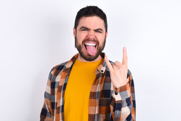Portrait of a crazy young caucasian man wearing plaid shirt over white background showing tongue horns up gesture, expressing excitement of being on concert of band.