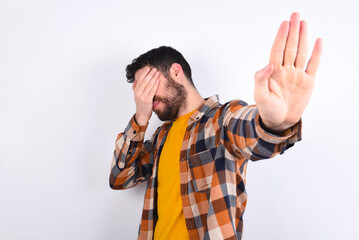 young caucasian man wearing plaid shirt over white background covers eyes with palm and doing stop gesture, tries to hide. Don't look at me, I don't want to see, feels ashamed or scared.