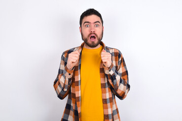 Portrait of desperate and shocked young caucasian man wearing plaid shirt over white background looking panic, holding hands near face, with mouth wide open.