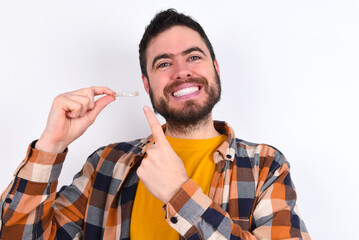 young caucasian man wearing plaid shirt over white background holding an invisible aligner and...