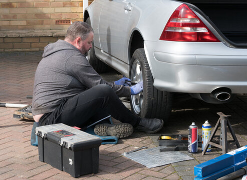 A male mechanic sits on the ground working on a silver sports car wheel.Many tools and parts lie on floor around him