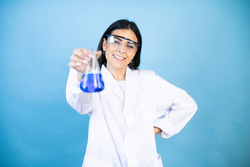 Young brunette woman wearing scientist uniform holding test tube over isolated blue background with a happy face standing and smiling with a confident smile showing teeth