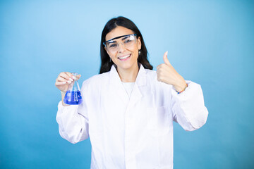 Young brunette woman wearing scientist uniform holding test tube over isolated blue background success sign doing positive gesture with hand, thumb up smiling and happy. cheerful expression