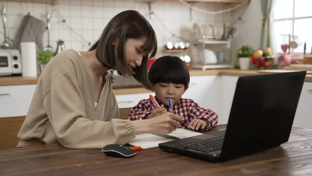 Asian mother supporting her son to write schoolwork at dining table while he is learning online from home with a laptop