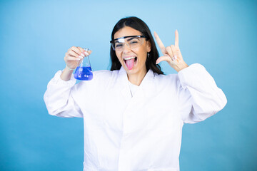 Young brunette woman wearing scientist uniform holding test tube over isolated blue background shouting with crazy expression doing rock symbol with hands up