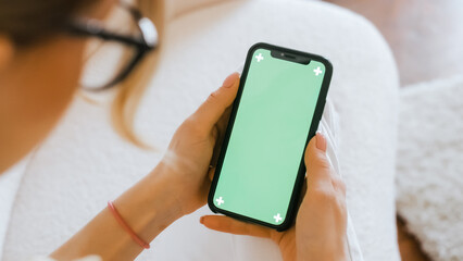 Young woman sitting on sofa at home and uses smartphone with green screen.